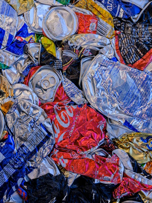 A close-up view of a large pile of crushed and crumpled aluminum cans, predominantly silver in color, with some featuring recognizable branding such as a red Coca-Cola logo and other colourful labels.