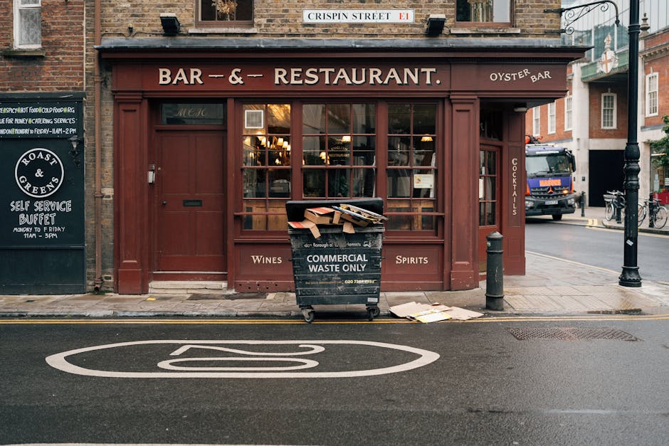 A black commercial waste bin positioned on the sidewalk in front of a red brick building housing a bar and restaurant. The bin has a sign indicating it is for 'Commercial Waste Only' and contains disc
